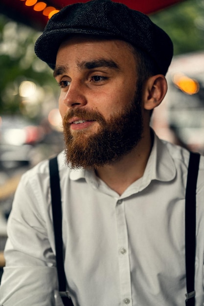 Joven barbudo en un café en la calle con una copa de vino. Chico romántico con una gorra de camisa blanca y tirantes en la ciudad. Peaky Blinders. antigua moda retro.