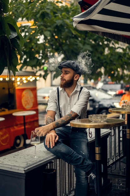 Joven barbudo en un café en la calle con una copa de vino. Chico romántico con una gorra de camisa blanca y tirantes en la ciudad. Peaky Blinders. antigua moda retro.