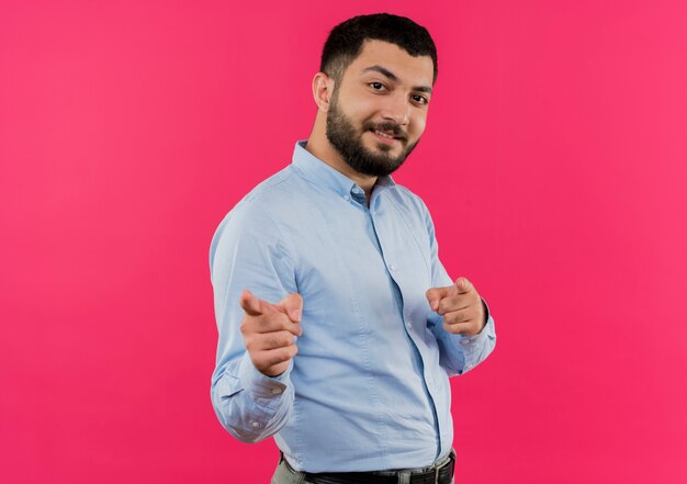 Joven barbudo con camisa azul apuntando con el dedo índice a la cámara sonriendo feliz y positivo