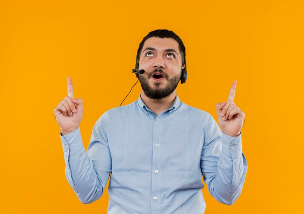 Joven barbudo con camisa azul con auriculares con micrófono apuntando hacia arriba con los dedos índices con cara feliz