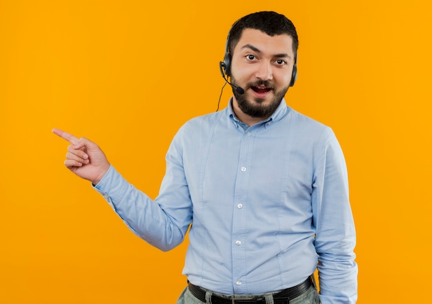 Joven barbudo con camisa azul con auriculares con micrófono apuntando con el dedo índice al lado sonriendo