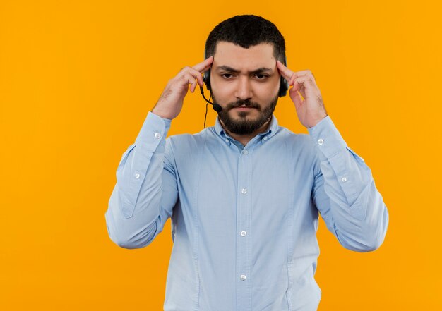 Joven barbudo con camisa azul con auriculares con micrófono apuntando sus sienes concentrándose en una tarea difícil