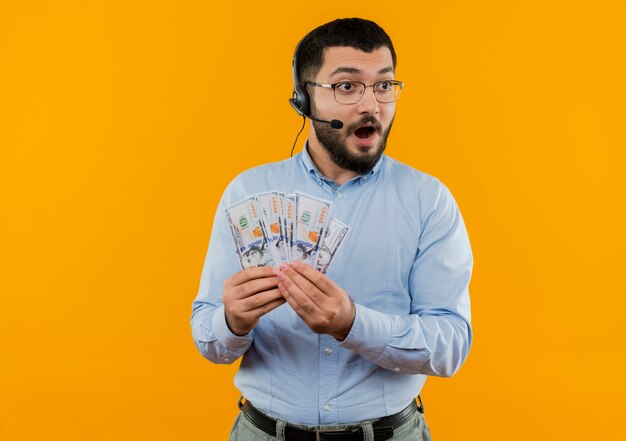 Joven barbudo con camisa azul con auriculares con micrófono mostrando dinero en efectivo feliz y sorprendido