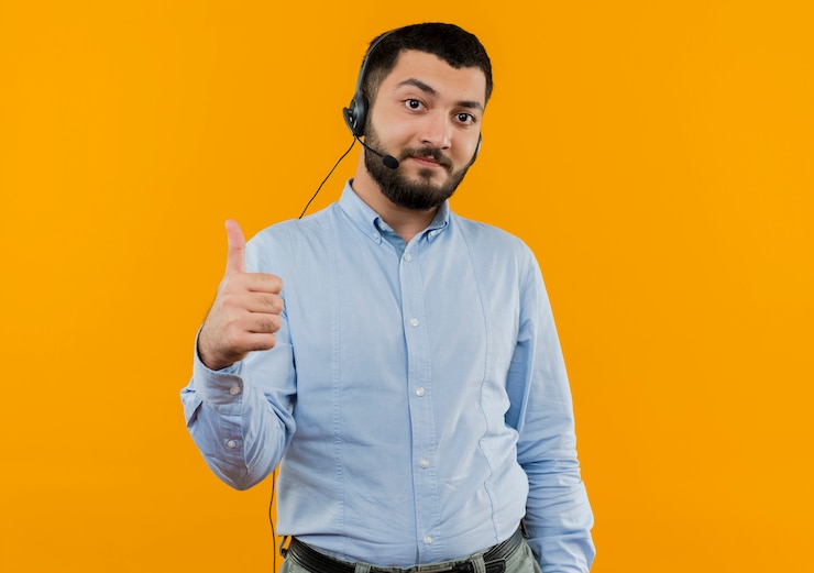 Foto gratuito joven barbudo con camisa azul con auriculares con micrófono sonriendo mostrando los pulgares para arriba