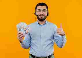 Foto gratuita joven barbudo con camisa azul con auriculares con micrófono sosteniendo dinero en efectivo mostrando los pulgares para arriba sonriendo con cara feliz