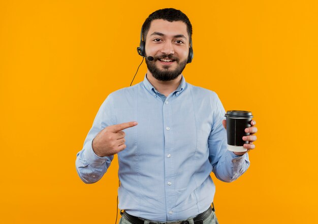 Joven barbudo con camisa azul con auriculares con micrófono sosteniendo la taza de café apuntando con el dedo índice sonriendo