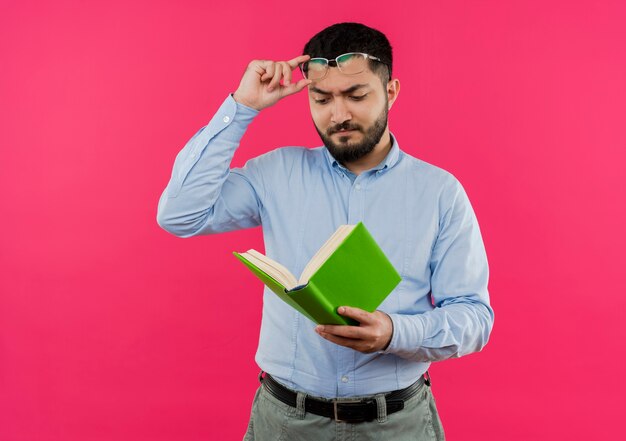 Joven barbudo con camisa azul mirando el libro en sus manos quitándose las gafas