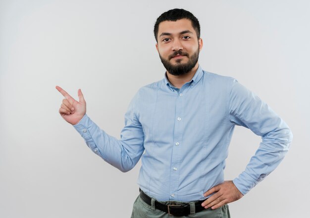 Joven barbudo en camisa azul pointign con el dedo índice hacia el lado sonriendo confiado de pie sobre la pared blanca