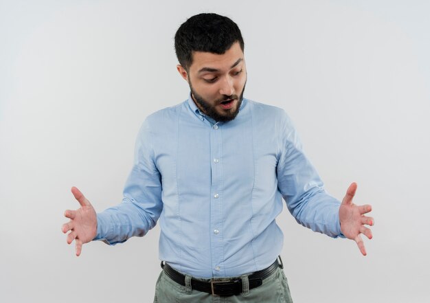 Joven barbudo con camisa azul que muestra un gesto de gran tamaño con las manos sorprendidas de pie sobre la pared blanca