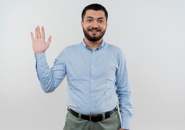 Joven barbudo con camisa azul saludando con la mano sonriendo