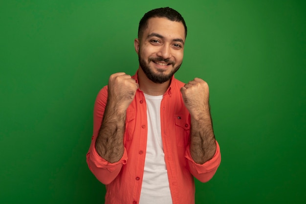 Joven barbudo en camisa naranja apretando los puños feliz y emocionado regocijándose de su éxito de pie sobre la pared verde