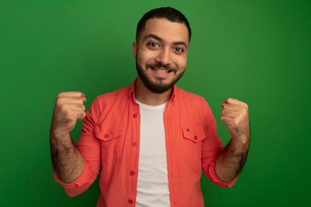 Joven barbudo en camisa naranja apretando los puños feliz y emocionado regocijándose de su éxito de pie sobre la pared verde