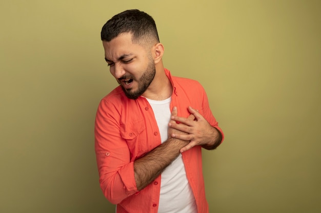 Foto gratuita joven barbudo con camisa naranja cogidos de la mano en el pecho sintiendo dolor de pie sobre una pared de luz