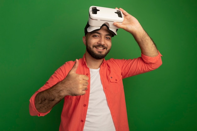 Joven barbudo en camisa naranja con gafas de realidad virtual en la cabeza mostrando thums sonriendo alegremente de pie sobre la pared verde