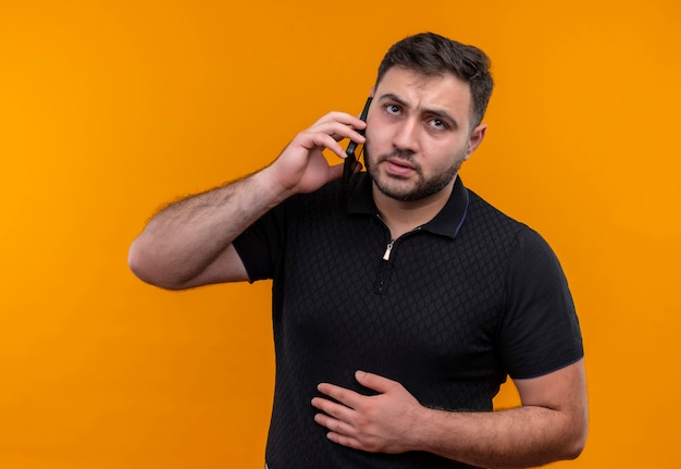 Joven barbudo con camisa negra que parece confundido y muy ansioso mientras habla por teléfono móvil