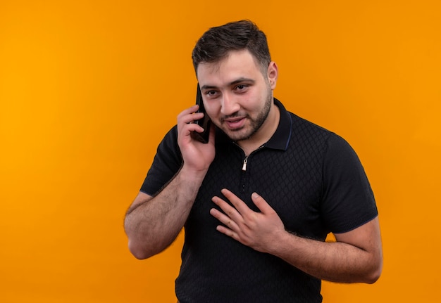 Joven barbudo en camisa negra sonriendo mientras habla por teléfono móvil