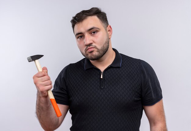 Joven barbudo con camisa negra sosteniendo un martillo mirando a la cámara con cara seria