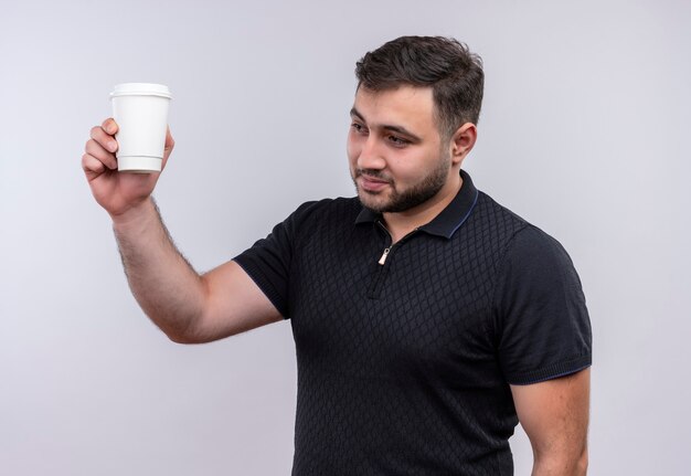 Joven barbudo con camisa negra sosteniendo la taza de café levantando la mano sonriendo confiado