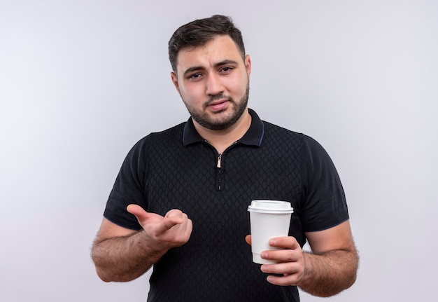 Joven barbudo con camisa negra sosteniendo la taza de café mirando a la cámara disgustado gesticulando con la mano