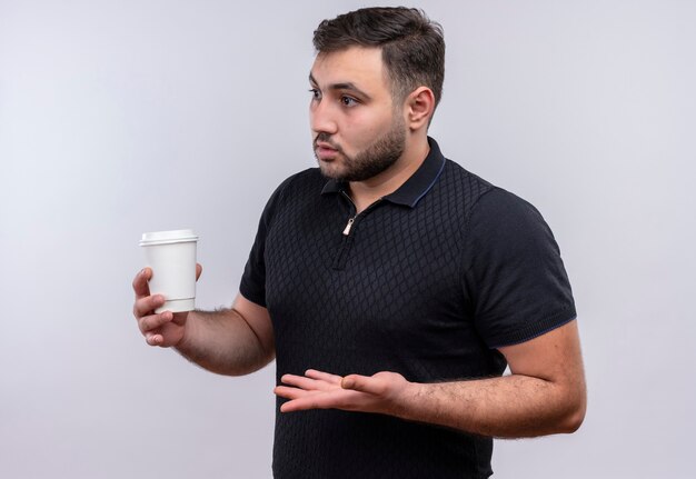 Joven barbudo con camisa negra sosteniendo la taza de café mirando a un lado disgustado gesticulando con la mano