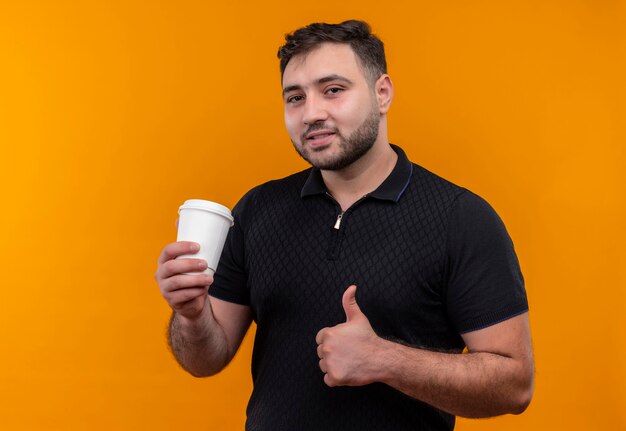 Joven barbudo con camisa negra sosteniendo una taza de café mostrando los pulgares para arriba sonriendo