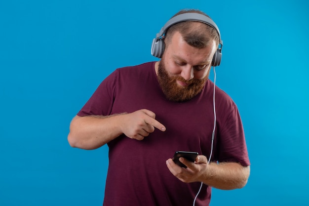 Joven barbudo en camiseta marrón con auriculares sosteniendo teléfono móvil buscando canción favorita