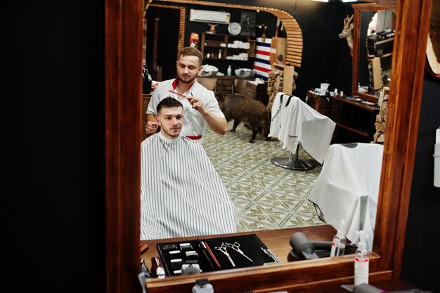 Joven barbudo cortándose el pelo con un peluquero mientras se sienta en una silla en la barbería
