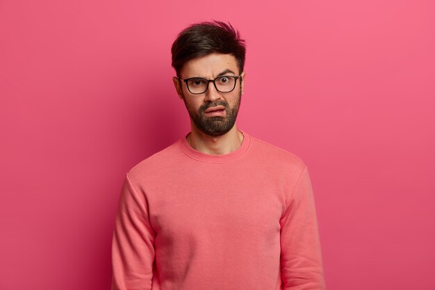 El joven barbudo descontento tiene una expresión de disgusto disgustado, reacciona ante algo desagradable, frunce el ceño, usa gafas y un jersey, se para en el interior contra una pared rosada. Concepto de emociones