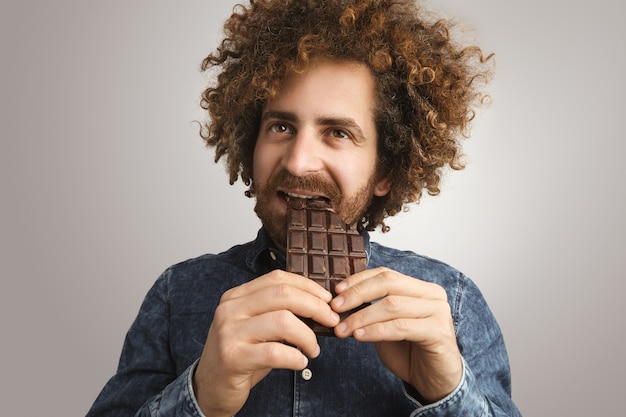 Foto gratuita joven barbudo feliz con piel sana y cabello rizado muerde soñadoramente una barra de chocolate orgánico recién horneado con el costado de la boca, sosteniendo la barra con ambas manos con cuidado