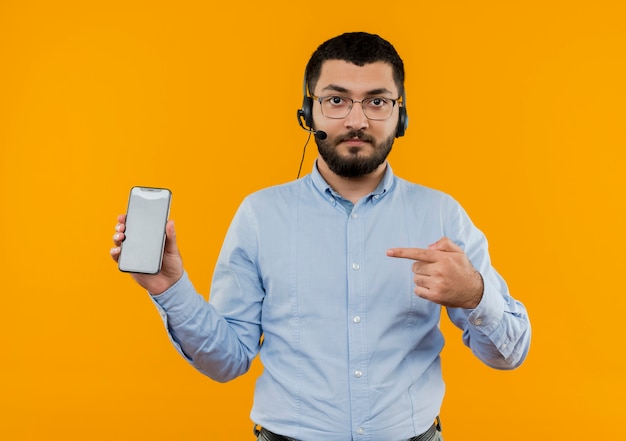 Joven barbudo con gafas y camisa azul con auriculares con micrófono mostrando smartphone apuntando con el dedo índice con cara seria