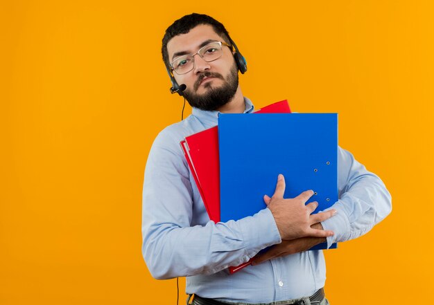 Joven barbudo con gafas y camisa azul con auriculares con micrófono sosteniendo carpetas con cara seria