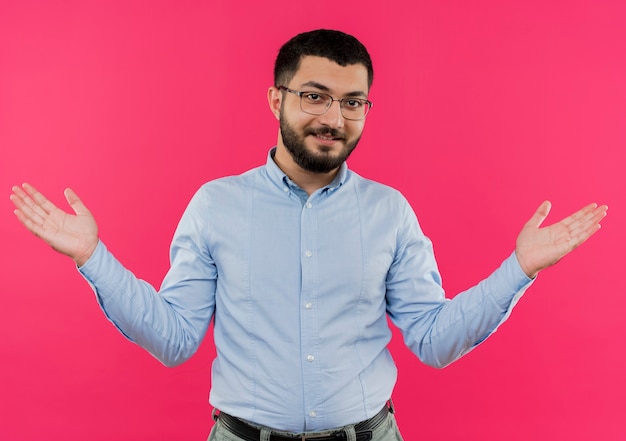 Joven barbudo con gafas y camisa azul extendiendo los brazos hacia los lados sonriendo con cara feliz