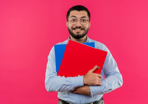 Joven barbudo con gafas y camisa azul sosteniendo carpetas sonriendo alegremente