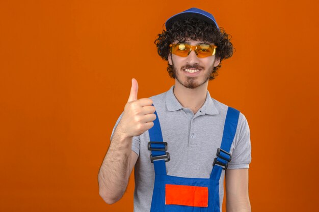 Joven barbudo guapo constructor con gafas y gorro de uniforme de construcción sonriendo mostrando el pulgar hacia arriba sobre la pared naranja aislada