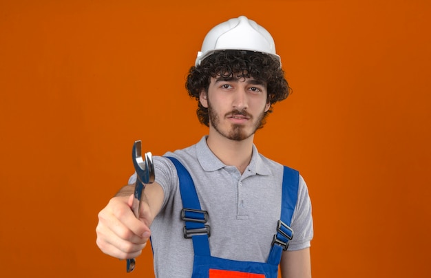 Foto gratuita joven barbudo guapo constructor vistiendo uniforme de construcción y casco de seguridad estirando llave con cara seria sobre pared naranja aislada