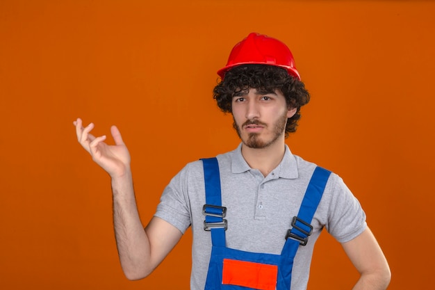 Joven barbudo guapo constructor vistiendo uniforme de construcción y casco de seguridad haciendo un gesto confuso con la expresión de la mano levantada como pregunta sobre pared naranja aislada