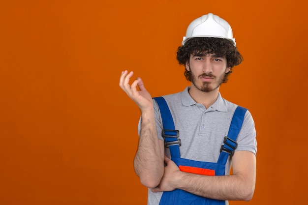 Joven barbudo guapo constructor vistiendo uniforme de construcción y casco de seguridad haciendo gesto confuso con la mano y la expresión como pregunta sobre la pared naranja aislada