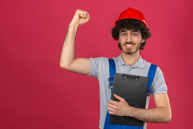 Joven barbudo guapo constructor vistiendo uniforme de construcción y casco de seguridad con portapapeles levantando el puño con cara feliz después del concepto de ganador de la victoria sobre la pared rosada aislada