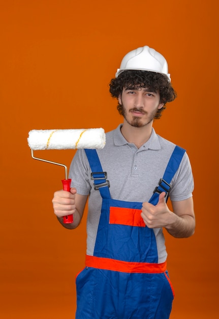 Joven barbudo guapo constructor vistiendo uniforme de construcción y casco de seguridad con rodillo de pintura mostrando el pulgar hacia arriba seguro de sí mismo sobre la pared naranja aislada