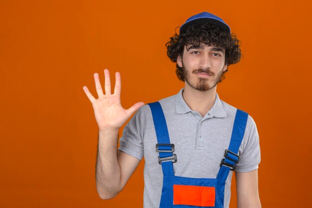 Joven barbudo guapo constructor vistiendo uniforme de construcción y gorra mostrando la palma abierta número cinco mientras sonríe amigable sobre pared naranja aislada