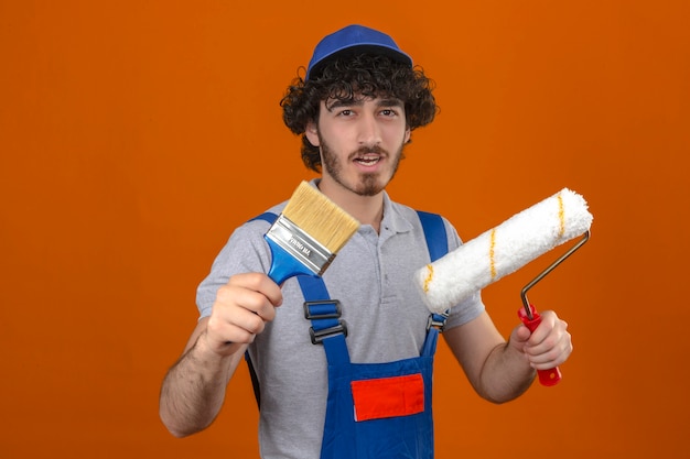 Joven barbudo guapo constructor vistiendo uniforme de construcción y gorra con rodillo de pintura y pincel sonriendo sobre pared naranja aislada