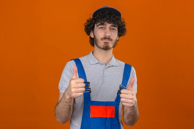 Joven barbudo guapo constructor vistiendo uniforme de construcción y gorra sonriendo mostrando los pulgares hacia arriba sobre la pared naranja aislada