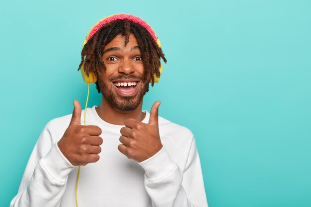 El joven barbudo se siente feliz, levanta dos pulgares, hace gestos similares, disfruta de su música favorita, se viste con ropa de diario