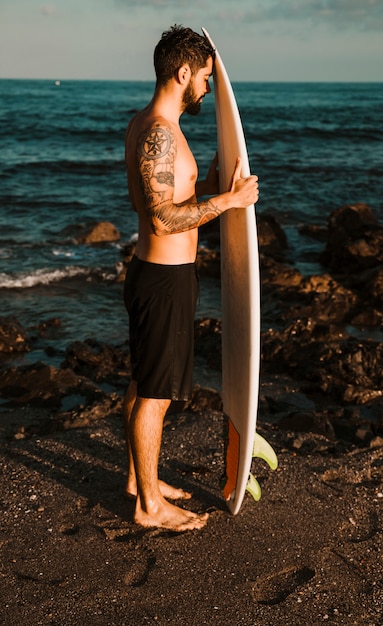 Joven barbudo con tabla de surf en la costa cerca del agua