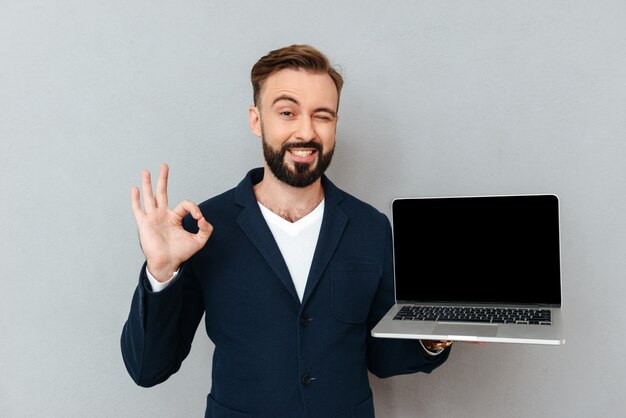 Joven barbudo en traje mirando la cámara mientras sostiene la computadora portátil aislada