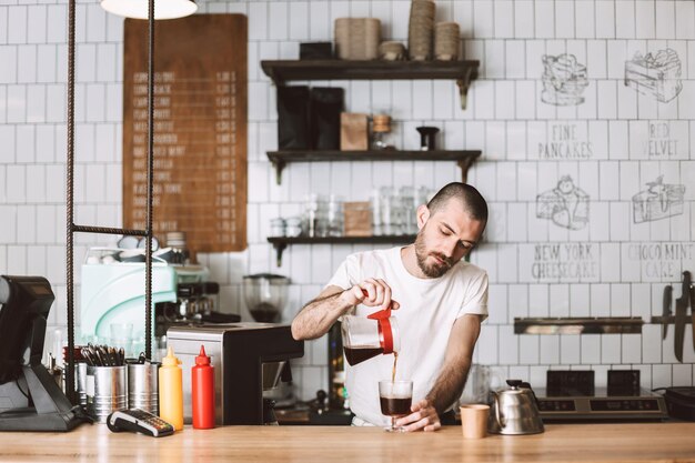 Joven barista parado en el mostrador del bar y vertiendo soñadoramente café de filtro en un vaso mientras trabaja en un café