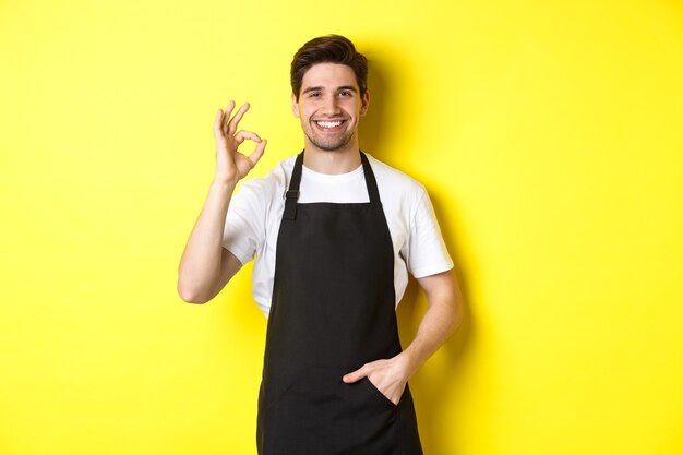Joven barista sonriente en delantal negro mostrando el signo bien, recomendando cafetería o restaurante, de pie sobre fondo amarillo