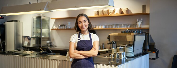Foto gratuito un joven barista sonriente con delantal trabajando en una cafetería parado cerca del mostrador con una máquina de café