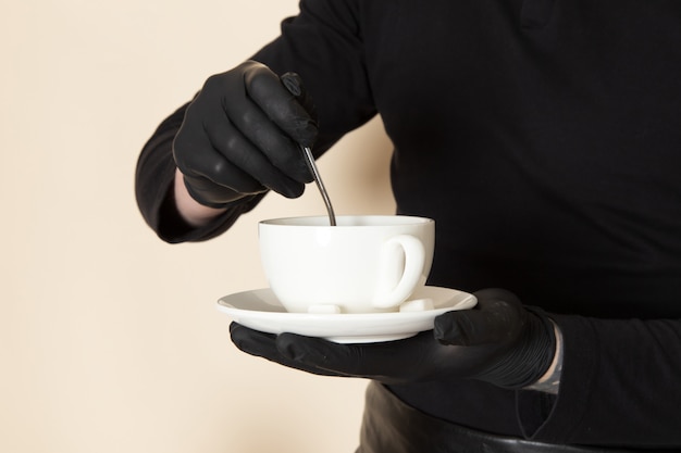 Joven barista en traje de trabajo negro con ingredientes y equipo de café semillas de café marrón con máscara estéril negra sobre blanco
