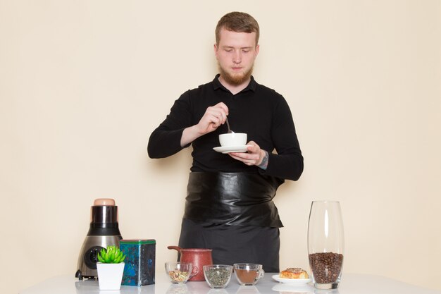 Joven barista en traje de trabajo negro con ingredientes y equipo de café semillas de café marrón sobre blanco